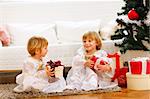 Two twins girl sitting with presents near Christmas tree