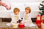 Two twins girls sitting with presents near Christmas tree