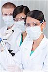 A female medical or scientific researcher or doctor using a pipette and test tube of clear liquid in a laboratory with her team of colleagues out of focus behind her.