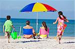 Rear view of a happy family of mother & father, parents daughter & son children having fun in deckchairs under an umbrella on a sunny beach