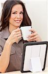 Beautiful, smiling, young brunette woman at home at a table using her tablet computer drinking a mug of tea or coffee