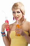 Young pretty girl with color make up hairstyle and color dress drinking from colored bottle some drink, she looks in to the lens, takes the rend bottle with right hand and drinks the orange drink