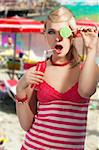 Young pretty girl with color make up hairstyle and color dress drinking from colored bottle some drink, she looks in to the lens with open mouth, covers her left eye with lollipop and accostes the the red bottle to her lips.