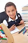 A beautiful young mixed race African American girl writing or drawing in a school classroom surrounded by books and an abacus