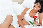 Portrait d'une femme souriante tenant une rose sur son lit dans une chambre lumineuse