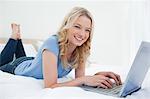 Une femme est couchée sur le lit en souriant et en regardant vers l'avant car elle utilise son ordinateur portable.