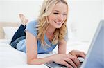 Une femme sourit comme elle tape loin sur son ordinateur portable, en position couchée sur le lit.