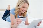 Une femme allongée sur le lit utilise son Tablet PC et la carte de crédit pour acheter des objets, elle sourit.