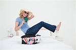 A woman is laughing as she wears glasses and hat, as she balances on her suitcase on the bed.