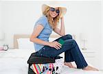 A woman sitting on her suitcase on the bed, with her passport in hand and wearing glasses and a hat.