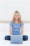 A woman sitting on a bed, with her laptop in front of her as she smiles and looks straight ahead.