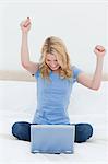 A woman is celebrating with arms in the air as she looks at her laptop while sitting on the bed.