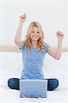A woman is looking forward with a smile as she celebrates on the bed in front of her laptop.