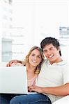 A man and woman smiling use the laptop while sitting on the couch together.
