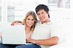 A smiling man and woman looking in front of them as they use the laptop on the couch.