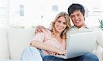 A man and woman sit on the couch together smiling as they use the laptop and look in front of them.
