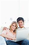 A man and woman sit embraced as they use the laptop while smiling and looking straight ahead.