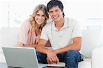 A man and woman smiling and looking ahead, as they sit on the couch with a laptop in front of them.