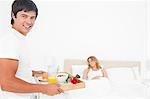 The woman is asleep as the man walks in with breakfast on a tray smiling.