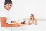 The man brings the woman breakfast in bed while both are smiling.