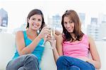 Two young smiling women sitting on a sofa while clinking glasses of champagne