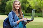 Young happy girl using her laptop while carrying her backpack and standing in a park