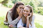 Young woman using her mobile phone while being told a secret by her friend