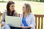 Young happy girls looking at each other while sitting on a bench and holding a laptop