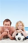 Playful young father and son lying on the beach with a football