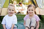 A smiling brother and sister sit inside of the tent with their parents outside