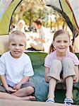 A smiling brother and sister sit inside of the tent as their parents sit outside talking