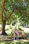 A smiling girl and her mom sit on a bench with a large tree providing shade