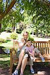 A smiling mom and girl sit together on a bench in the park near a large tree
