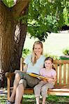 A smiling mom and her daughter smile and look at the camera with a book in hand and sitting on the park bench