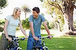 A smiling couple sitting on their bikes in the park looking at one another