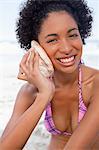 Young smiling woman in beachwear looking at the camera while listening to a shell