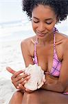 Young woman in beachwear looking at a shell while sitting down on the beach