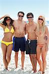 Two men and two women in swimsuits smaile as they put their arms around each other on the beach