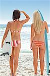 Woman in a bikini looking over the sea with her friend as they both hold surfboards