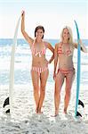 Two women in bikinis smiling happily as they stand next to perched surfboards on the beach