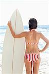 Frau im Bikini ein Surfbrett gedrückt halten, während sie einerseits auf ihre Hüfte beruht und auf das Meer schaut