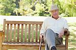 Man holding a cane and sitting on a bench while he looks ahead of him in a park