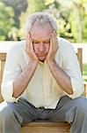 Man looking towards the ground as he rests his head between his hands while he sits on a bench in a park