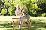 Man plays a guitar while standing with a leg raised in front of a bench in a park