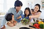 A family all help and watch to make and toss a salad together in the kitchen