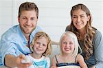 The smiling family sit on the couch together as the dad changes the tv channel