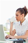 Frizzy haired woman drinking a coffee while tapping on her laptop in a bright office