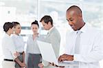 Smiling young salesman using laptop with colleagues behind him