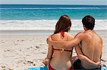Rear view of a young tanned couple sitting on beach towel in front of the sea