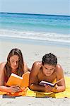 Tanned couple reading a book while lying on the beach with the sea in background